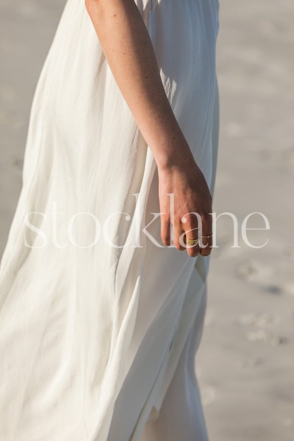 Vertical stock photo of a barefoot woman at the beach, wearing a white dress.