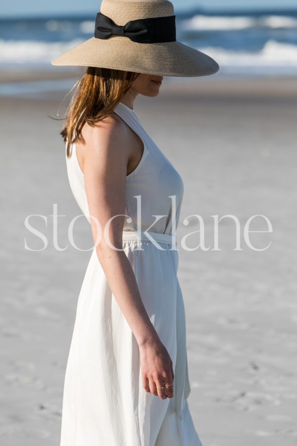 Vertical stock photo of a woman wearing a white dress and a hat, walking on the beach.