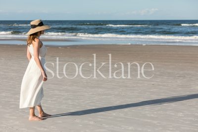 Horizontal stock photo of a woman wearing a white dress and a hat, walking on the beach.