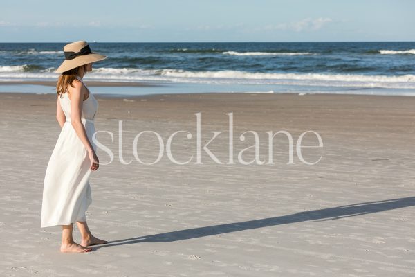 Horizontal stock photo of a woman wearing a white dress and a hat, walking on the beach.