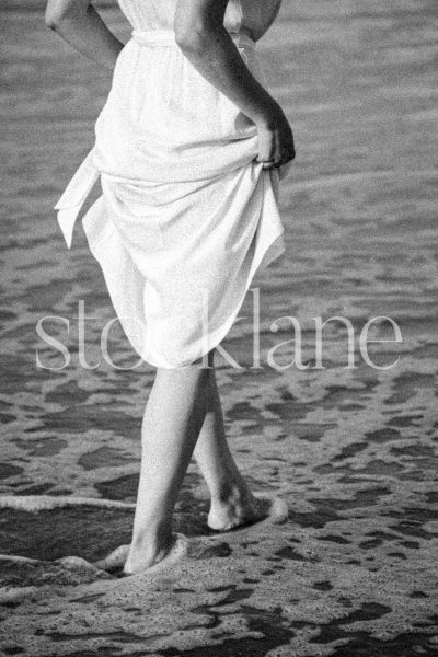 Vertical black and white stock photo of a woman wearing a white dress walking into the water.