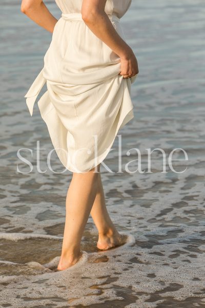 Vertical stock photo of a woman wearing a white dress walking into the water.