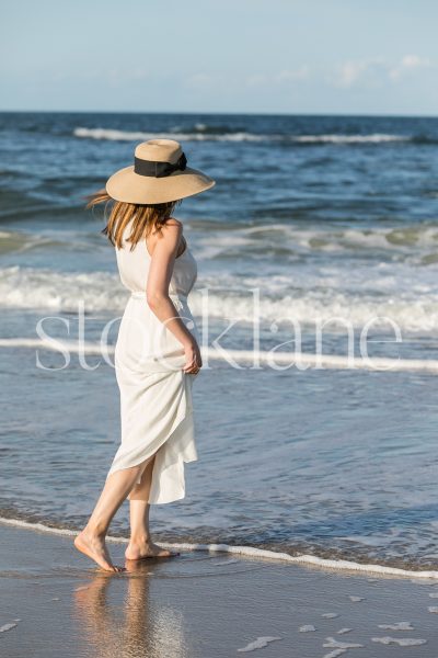 Vertical stock photo of a woman wearing a white dress and a hat, walking on the beach.