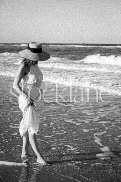 Vertical black and white stock photo of a woman wearing a white dress walking into the water.