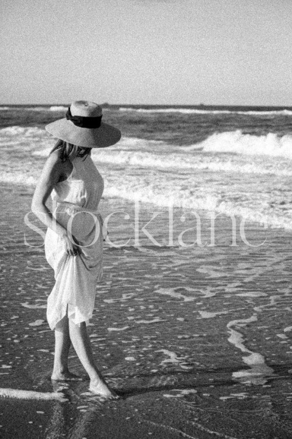 Vertical black and white stock photo of a woman wearing a white dress walking into the water.