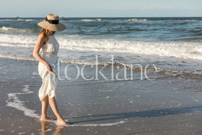 Horizontal stock photo of a woman wearing a white dress and a hat, walking on the beach, into the water.