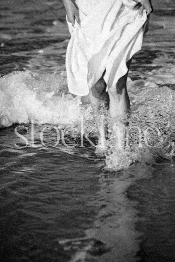 Vertical black and white photo of a woman wearing a white dress splashing in the water at the beach.