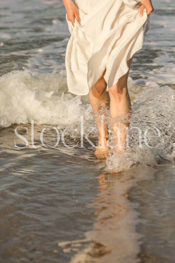 Vertical photo of a woman wearing a white dress splashing in the water at the beach.