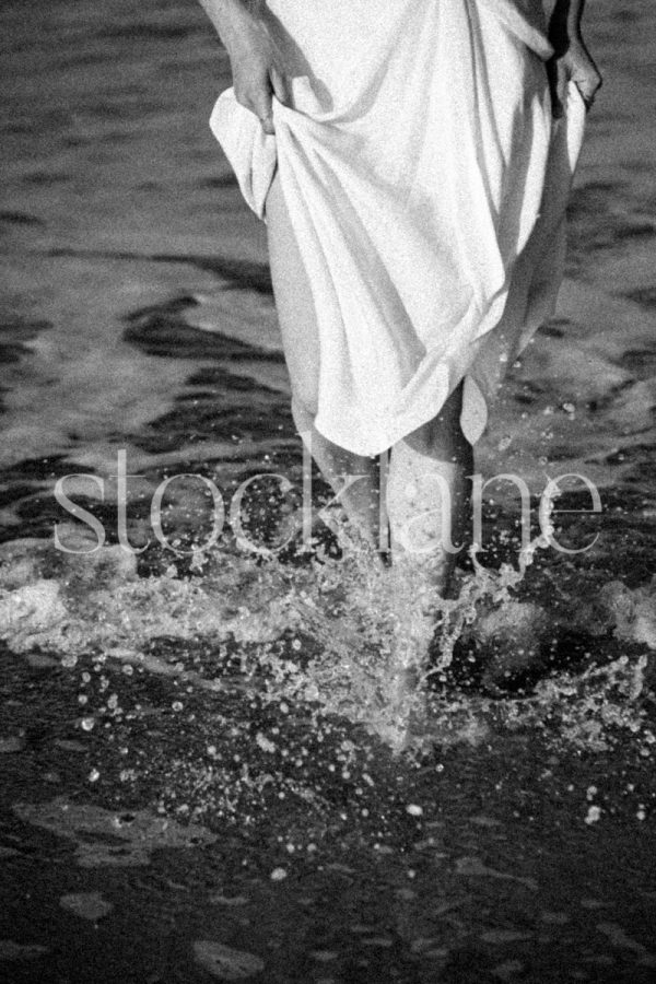 Vertical black and white photo of a woman wearing a white dress splashing in the water at the beach.