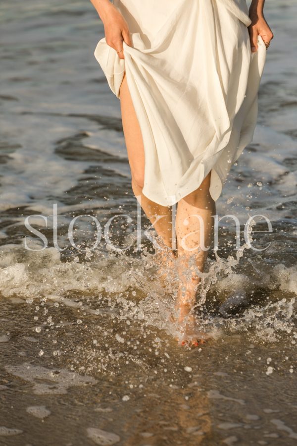 Vertical photo of a woman wearing a white dress splashing in the water at the beach.