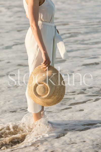 Vertical stock photo of a woman at the beach, standing in the surf, wearing a white dress and holding a straw hat by her side.