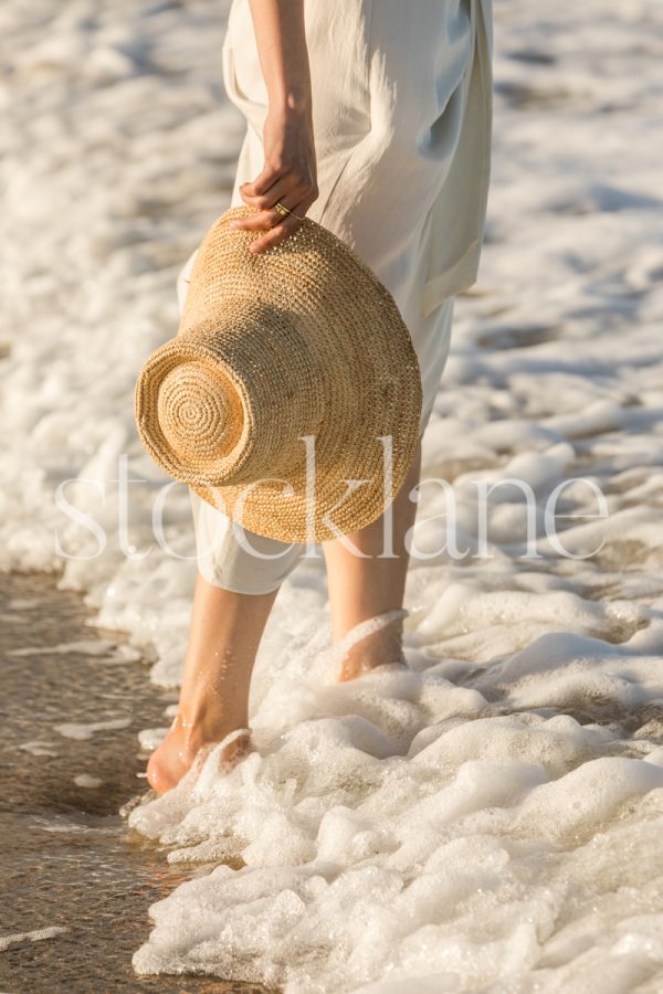 Vertical stock photo of a woman at the beach, standing in the surf, wearing a white dress and holding a straw hat by her side.