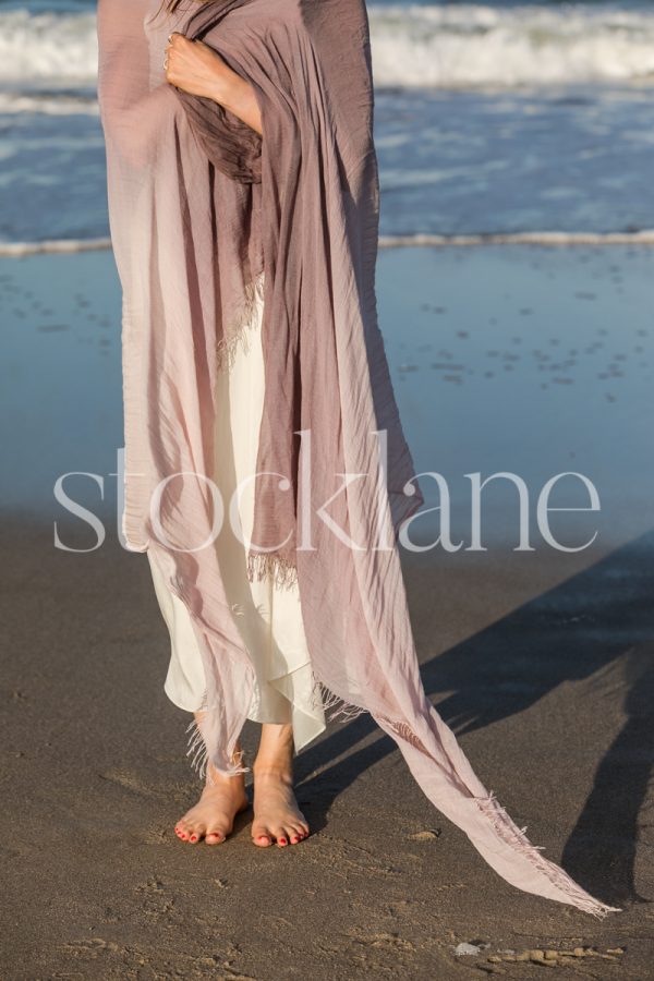 Vertical stock photo of a woman wrapped in a purple shawl, barefoot, at the beach