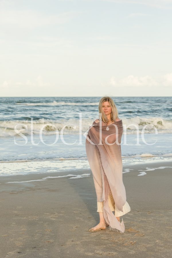 Vertical stock photo of a woman wrapped in a purple shawl, at the beach, standing in front of the water.