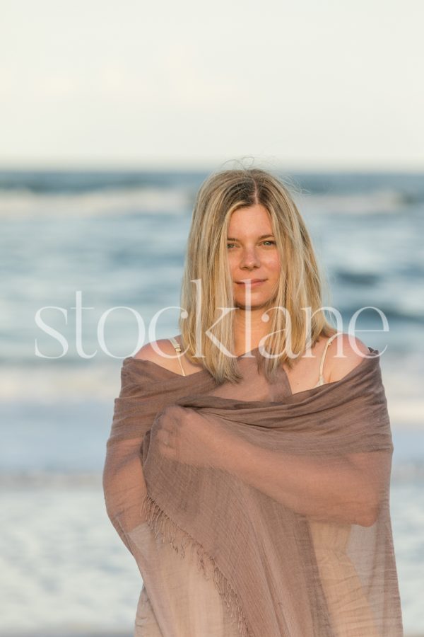 Vertical stock photo of a woman wrapped in a purple shawl, at the beach, standing in front of the water.