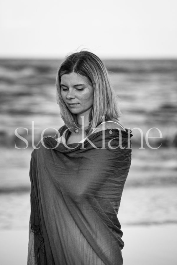 Vertical black and white photo of a woman wrapped in a shawl at the beach.