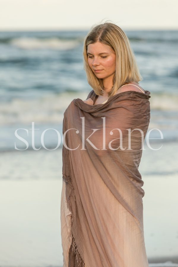 Vertical stock photo of a woman wrapped in a purple shawl, at the beach, standing in front of the water.