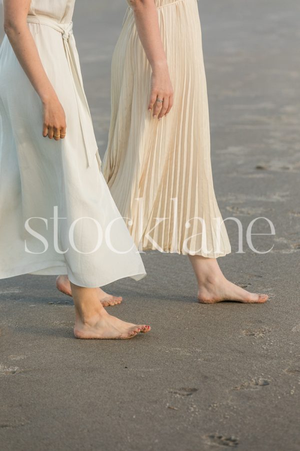 Vertical stock photo of two women in light colored dresses, walking side by side at the beach.