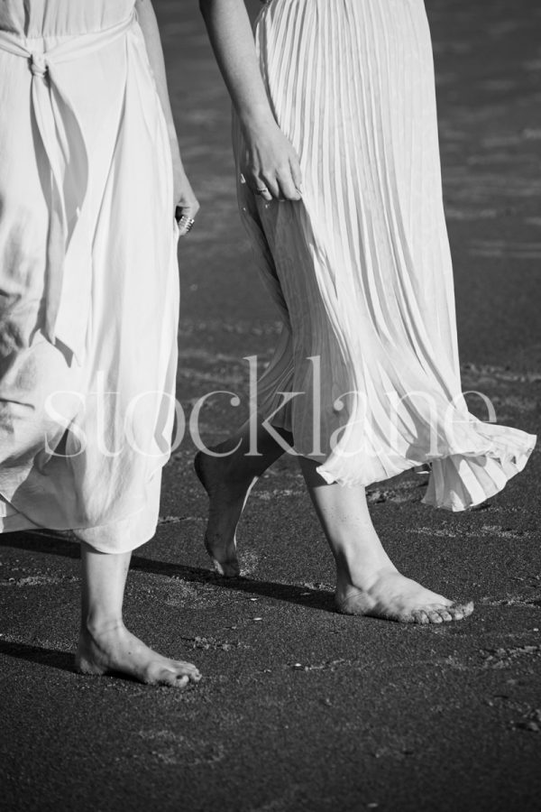 Vertical black and white stock photo of two women in light colored dresses, walking side by side at the beach.