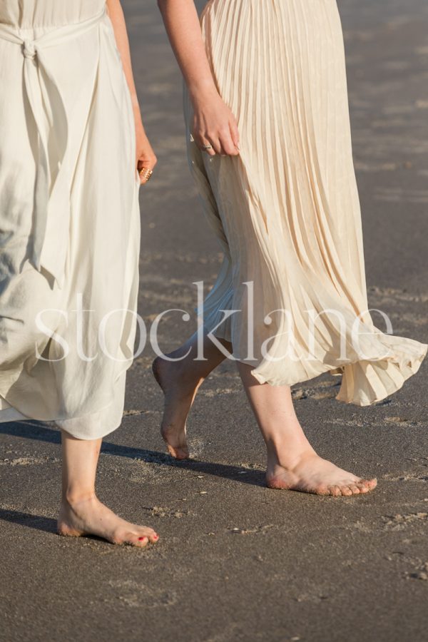 Vertical stock photo of two women in light colored dresses, walking side by side at the beach.