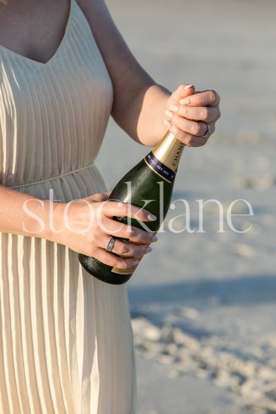 Vertical stock photo of a woman wearing a cream pleated dress, opening a bottle of champagne.