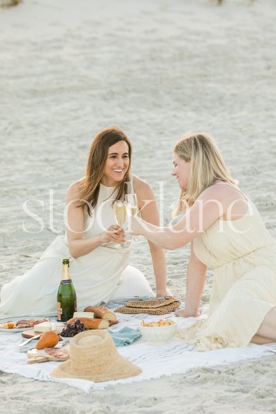 Vertical stock photo of two women, wearing light colored dresses, having a picnic at the beach.