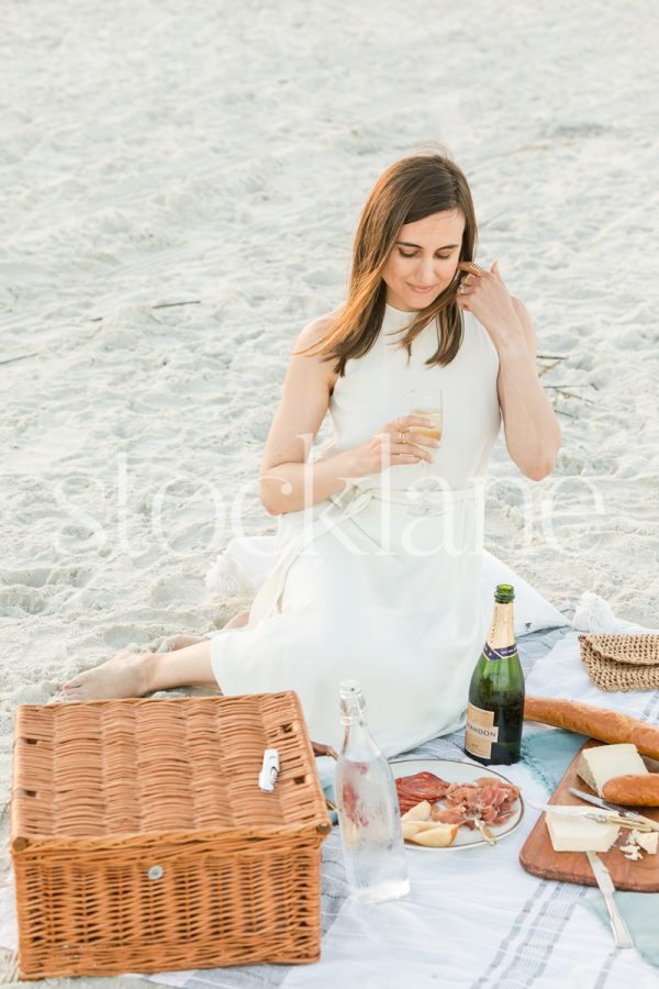 Vertical stock photo of a woman holding a glass of champagne while having a picnic at the beach.