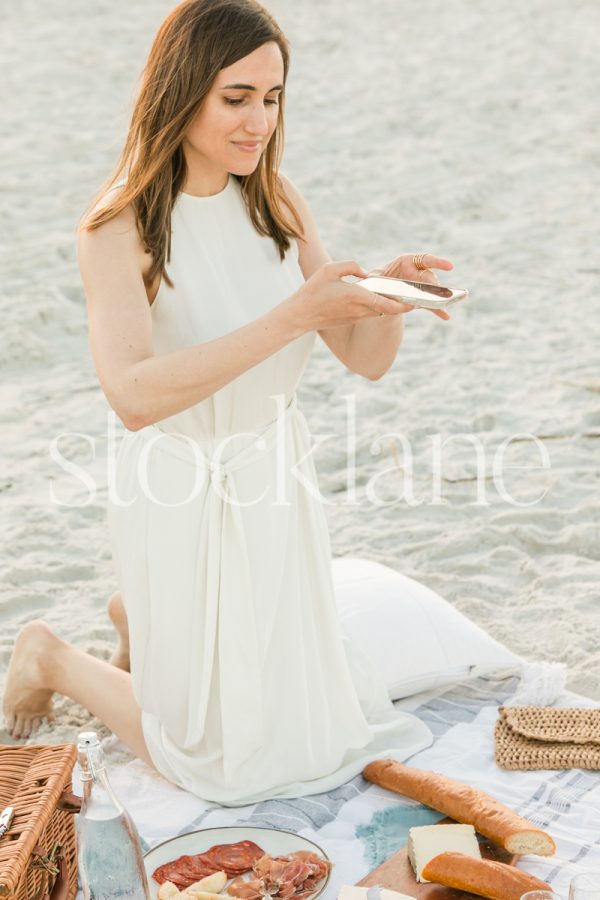 Vertical stock photo of a woman wearing a white dress taking an overhead photo of a picnic at the beach, with her phone.