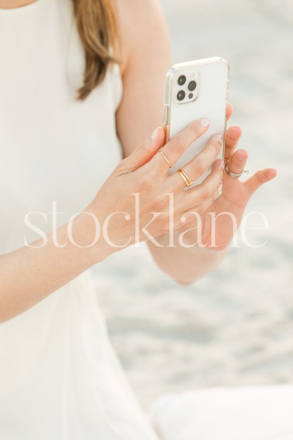 Vertical stock photo of a woman wearing a white dress taking a photo with her phone.