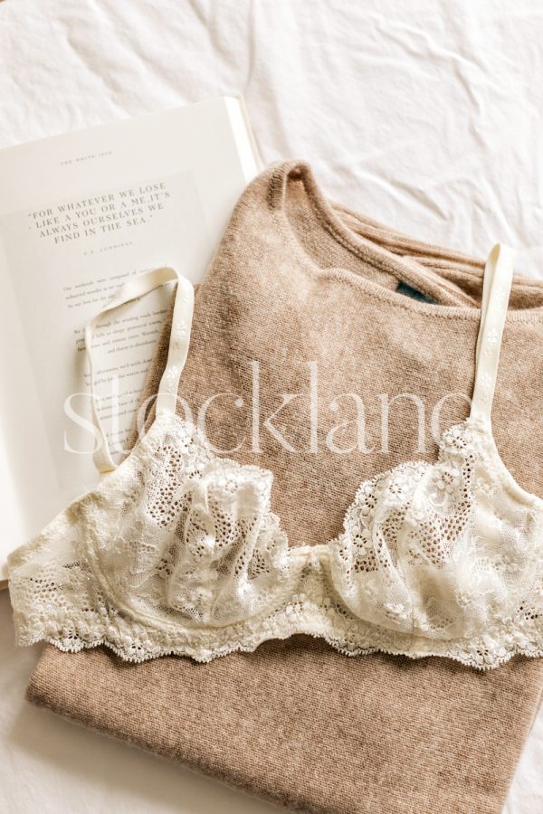 Vertical stock photo of a book, a sweater and a lace bra on a white bed sheet.