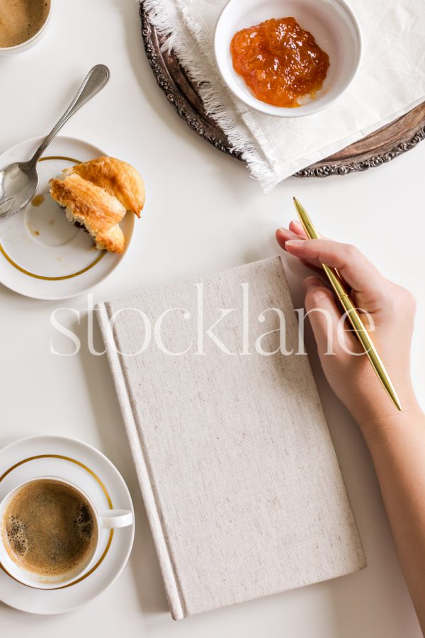 Vertical stock photo of a woman's hand holding a gold pen next to a linen-bound notebook, on a table with coffee and other food.