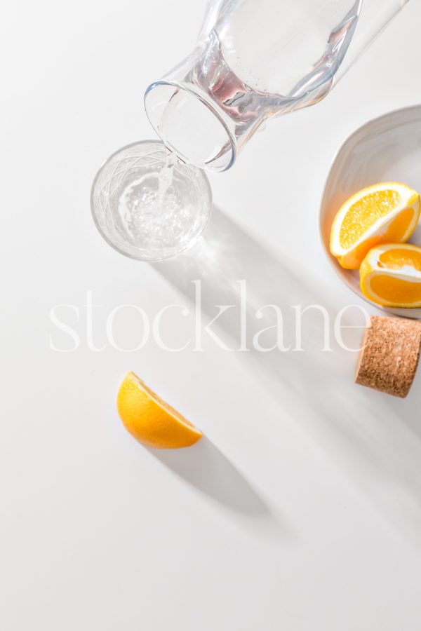 Vertical stock photo of a jar of water being poured into a glass, with sliced oranges around it.