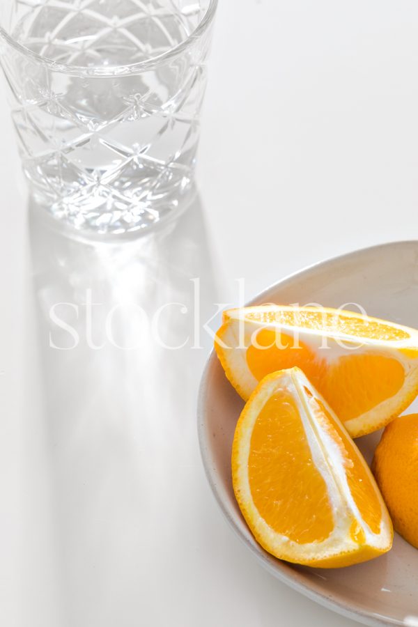 Vertical stock photo of a glass of water with light reflecting through it, next to a plate of sliced oranges.