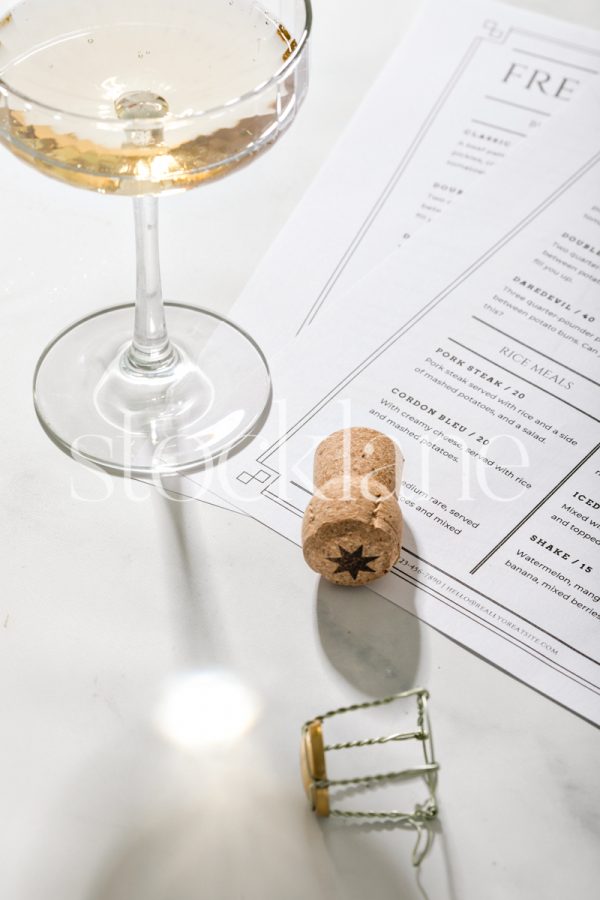 Vertical stock photo of a coupe glass of champagne next to a cork and restaurant menus.