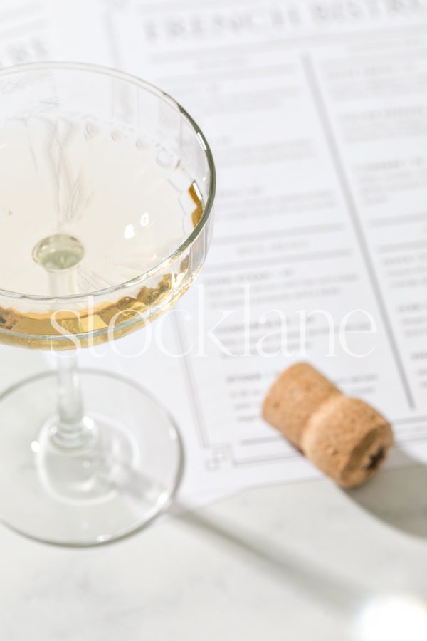 Vertical stock photo of a coupe glass of champagne next to a cork and restaurant menus.