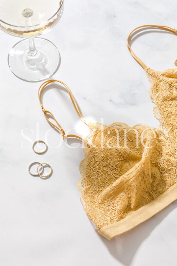 Vertical stock photo of a champagne glass, rings, and a yellow bralette on a marble surface.