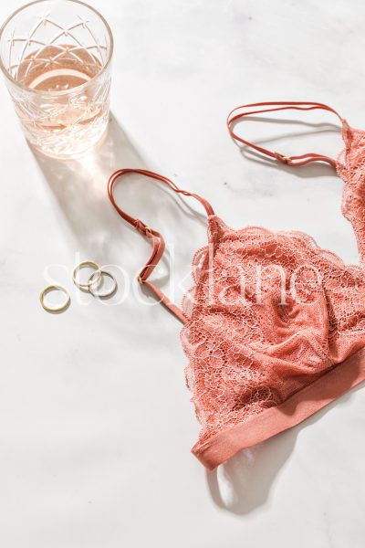 Vertical stock photo of a glass of rose wine, rings, and a pink bralette on a marble surface.