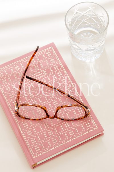 Vertical stock photo of a pink notebook, with tortoise glasses on top, and a glass of water, with sunlight coming through window blinds.