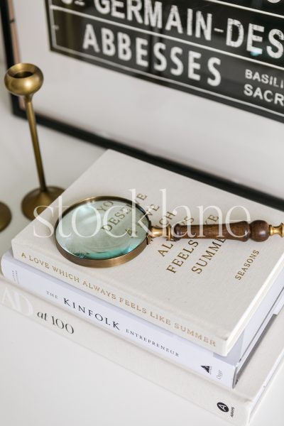 Vertical stock photo of an antique magnifying glass on top of a stack of books in neutral colors.