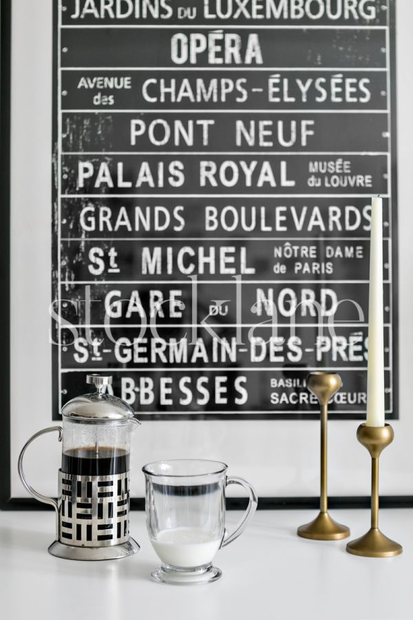 Vertical stock photo of a French press and a glass of milk on a white table top with candle holders and a print in the background.