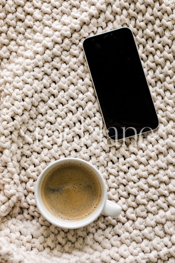 Vertical stock photo of a coffee cup and a phone on a neutral colored blanket.