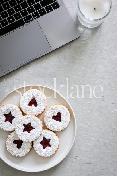 Vertical stock photo of a laptop, a plate of linzer cookies and a candle.