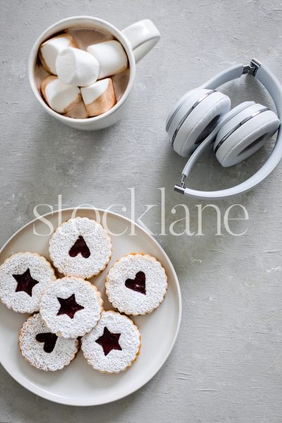 Vertical stock photo of a cup of hot cocoa, a plate of linzer cookies and headphones.