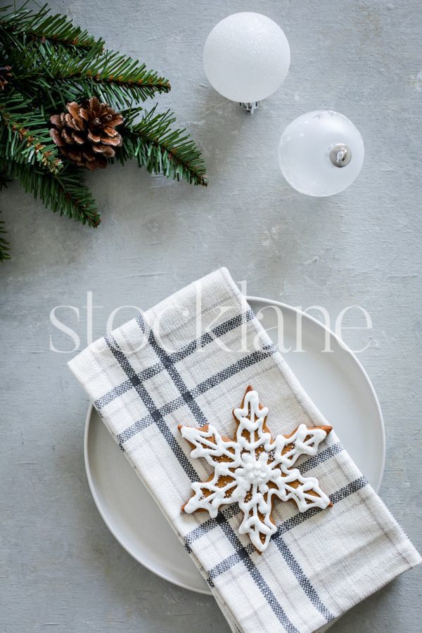 Vertical stock photo of a plate with a snowflake gingerbread cookie, with Christmas ornaments and greenery.