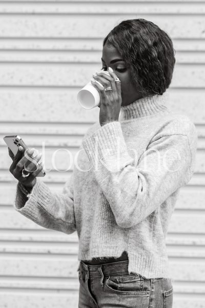 Vertical black and white photo of a woman drinking coffee while looking at her phone.