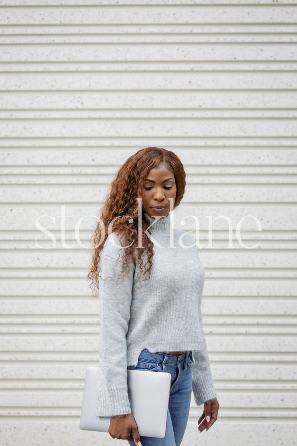 Vertical stock photo of a woman wearing a gray sweater and jeans, holding a laptop computer