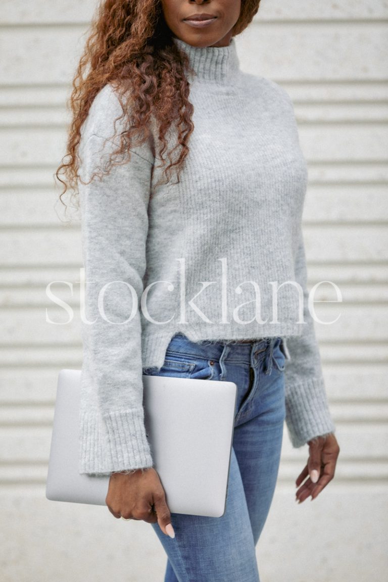 Vertical stock photo of a woman wearing a gray sweater and jeans, holding a laptop computer.