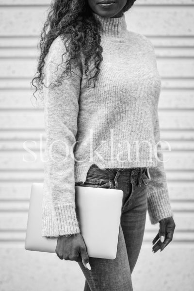 Vertical black and white stock photo of a woman wearing a gray sweater and jeans, holding a laptop computer.