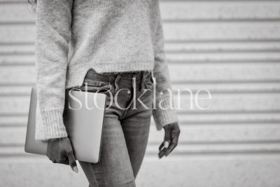 Horizontal black and white stock photo of a woman wearing a gray sweater and jeans, holding a laptop computer.