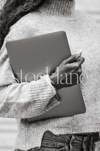 Vertical black and white stock photo of a woman wearing a gray sweater holding a laptop computer.